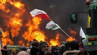 Rolnicy protestujący w Brukseli. W centralnym punkcie polska flaga, na tle płomieni ognia, obok widoczny jest ciągnik rolniczy.