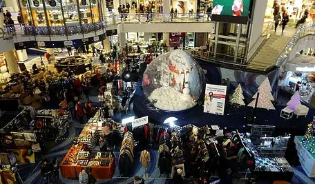 Świątecznie udekorowane centrum handlowe pełne klientów; widok z góry na stoiska z odzieżą, ozdoby bożonarodzeniowe oraz dużą śnieżną kulę na środku przestrzeni.