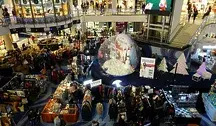 Świątecznie udekorowane centrum handlowe pełne klientów; widok z góry na stoiska z odzieżą, ozdoby bożonarodzeniowe oraz dużą śnieżną kulę na środku przestrzeni.