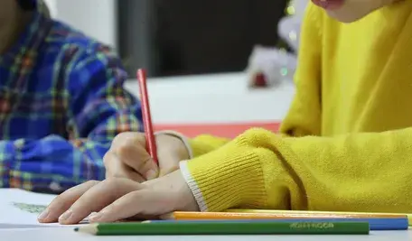 Dziecko w żółtym swetrze rysujące kolorową kredką przy stole, obok leżą inne ołówki i drugie dziecko w koszuli w kratę.