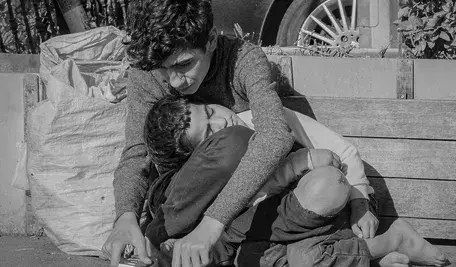 Dwoje dzieci siedzi na chodniku przy ławce; starsze obejmuje młodsze, które leży oparte o jego kolana, obok nich znajduje się torba z rzeczami osobistymi.