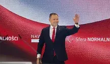 Prezydent Karol Nawrocki podczas wystąpienia publicznego na biało-czerwonym tle. Uśmiecha się i macha ręką w kierunku widowni.