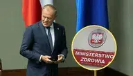 Donald Tusk w garniturze stoi w sali obrad obok biało-czerwonej i niebieskiej flagi; w rogu zdjęcia znajduje się okrągłe zbliżenie na tablicę z napisem ‘Ministerstwo Zdrowia’ i godłem Polski.