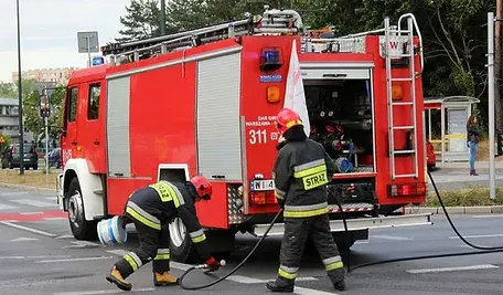 Strażacy gaszą pożar przy czerwonym wozie strażackim na skrzyżowaniu w mieście.