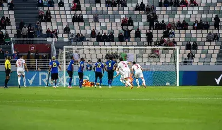 Piłkarz w białym stroju wykonuje rzut karny podczas meczu piłkarskiego, piłka wpada do bramki obok interweniującego bramkarza w pomarańczowym stroju, a zawodnicy drużyny w niebieskich koszulkach stoją w polu karnym; w tle trybuny stadionu z kibicami.