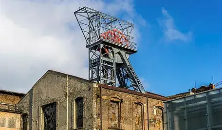 Wieża wyciągowa kopalni węgla z widocznym szybikiem i zabudowaniami przemysłowymi na tle błękitnego nieba.