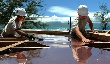 Dwóch pracowników budowlanych w kaskach ochronnych montuje przezroczystą płytę na drewnianej konstrukcji dachu, pracując na wysokości przy słonecznej pogodzie, z widokiem na morze w tle.