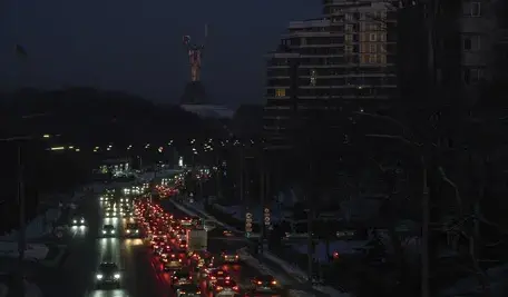 Zdjęcie nocne przedstawiające ulicę w centrum Kijowa podczas awarii zasilania. Ciemne sylwetki bloków mieszkalnych i biurowców odcinają się na tle nocnego nieba. Jedynym źródłem światła są smugi i punkty reflektorów przejeżdżających samochodów, podkreślające paraliż energetyczny miasta.