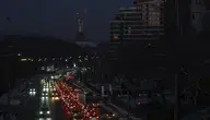 Zdjęcie nocne przedstawiające ulicę w centrum Kijowa podczas awarii zasilania. Ciemne sylwetki bloków mieszkalnych i biurowców odcinają się na tle nocnego nieba. Jedynym źródłem światła są smugi i punkty reflektorów przejeżdżających samochodów, podkreślające paraliż energetyczny miasta.