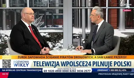 Telewizyjna rozmowa na żywo – dwóch mężczyzn w garniturach, aMarcin Wikło i prezes TK Bogdan Święczkowski siedzi przy stole w studiu, w tle zimowa sceneria miejska, na ekranie pasek informacyjny z hasłem o ściganiu piratów drogowych