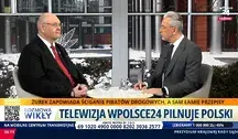 Telewizyjna rozmowa na żywo – dwóch mężczyzn w garniturach, aMarcin Wikło i prezes TK Bogdan Święczkowski siedzi przy stole w studiu, w tle zimowa sceneria miejska, na ekranie pasek informacyjny z hasłem o ściganiu piratów drogowych