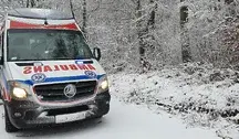 Karetka pogotowia ratunkowego na zaśnieżonej, leśnej drodze, jadąca na sygnale w zimowych warunkach.