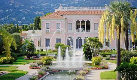 Elegancka willa w pastelowym różu otoczona zadbanym ogrodem z palmami, fontanną i alejkami, na tle zielonych wzgórz