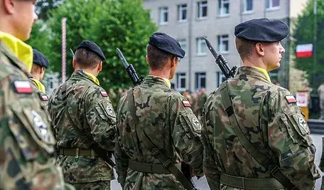 Żołnierze Wojska Polskiego w mundurach stoją w szyku tyłem do obiektywu, z karabinami na ramieniu, przed budynkiem jednostki wojskowej, widoczna polska flaga