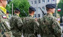 Żołnierze Wojska Polskiego w mundurach stoją w szyku tyłem do obiektywu, z karabinami na ramieniu, przed budynkiem jednostki wojskowej, widoczna polska flaga