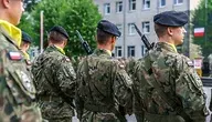 Żołnierze Wojska Polskiego w mundurach stoją w szyku tyłem do obiektywu, z karabinami na ramieniu, przed budynkiem jednostki wojskowej, widoczna polska flaga