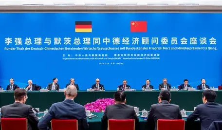 Kanclerz Niemiec Friedrich Merz i premier Chin Li Qiang siedzą przy długim stole konferencyjnym w Wielkiej Hali Ludowej w Pekinie podczas obrad okrągłego stołu niemiecko-chińskiego komitetu doradczego. W tle znajduje się wielka niebieska ściana z flagami Niemiec i Chin oraz napisem w języku niemieckim i chińskim. Przed politykami widać delegację biznesową oraz kompozycję z różowych kwiatów.