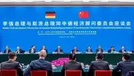 Kanclerz Niemiec Friedrich Merz i premier Chin Li Qiang siedzą przy długim stole konferencyjnym w Wielkiej Hali Ludowej w Pekinie podczas obrad okrągłego stołu niemiecko-chińskiego komitetu doradczego. W tle znajduje się wielka niebieska ściana z flagami Niemiec i Chin oraz napisem w języku niemieckim i chińskim. Przed politykami widać delegację biznesową oraz kompozycję z różowych kwiatów.