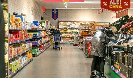 Dwoje ludzi chodzi po sklepie spożywczym między regałami wypełnionymi produktami.