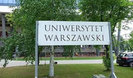 Biała tablica z napisem Uniwersytet Warszawski stojąca na tle drzew.