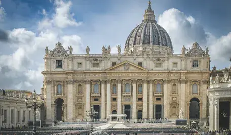Zdjęcie przedstawia Bazylikę św. Piotra w Watykanie pod błękitnym niebem z białymi chmurami. Na pierwszym planie widoczny jest wysoki, kamienny obelisk watykański, który przysłania centralną część fasady. W tle dominuje monumentalna kopuła bazyliki zwieńczona krzyżem. Na szczycie fasady widoczne są rzeźby apostołów oraz charakterystyczny zegar po lewej stronie. Na gzymsie widnieje łaciński napis: „IN HONOREM PRINCIPIS APOST PAVLVS V BVRGHESIVS ROMANVS PONT MAX AN MDCXII PONT VII”.