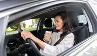 Kobieta siedzi w samochodzie i popija kawę. To srebrny sedan z kierownicą po prawej stronie. Kobieta jest młoda i uśmiechnięta