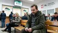 Fotografia przedstawia wnętrze poczekalni w polskim Powiatowym Urzędzie Pracy. Na pierwszym planie, na drewnianej ławce, siedzi zamyślony, brodaty mężczyzna w zielonej kurtce, patrzący na trzymany w dłoniach numerek. W tle widać inne oczekujące osoby oraz stanowiska obsługi z wyraźnymi napisami w języku polskim: „Czekaj A145”, „STANOWISKO 4”, „URZĄD PRACY” wraz z godłem Polski, co jednoznacznie wskazuje na kontekst poszukiwania zatrudnienia.