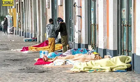 Koce i rzeczy osób bezdomnych rozłożone na ulicy. W tle widać dwóch mężczyzn