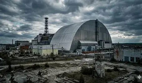 Szeroki kadr na Elektrownię Jądrową w Czarnobylu z lotu ptaka. Na pierwszym planie widoczny nowoczesny, stalowy arkusz Arki (New Safe Confinement) przykrywający zniszczony reaktor nr 4, obok charakterystyczny biało-czerwony komin wentylacyjny. Całość utrzymana w mrocznej, dramatycznej tonacji pod zachmurzonym, szarym niebem, podkreślająca postapokaliptyczny klimat strefy wykluczenia.