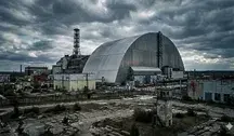 Szeroki kadr na Elektrownię Jądrową w Czarnobylu z lotu ptaka. Na pierwszym planie widoczny nowoczesny, stalowy arkusz Arki (New Safe Confinement) przykrywający zniszczony reaktor nr 4, obok charakterystyczny biało-czerwony komin wentylacyjny. Całość utrzymana w mrocznej, dramatycznej tonacji pod zachmurzonym, szarym niebem, podkreślająca postapokaliptyczny klimat strefy wykluczenia.