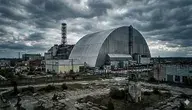 Szeroki kadr na Elektrownię Jądrową w Czarnobylu z lotu ptaka. Na pierwszym planie widoczny nowoczesny, stalowy arkusz Arki (New Safe Confinement) przykrywający zniszczony reaktor nr 4, obok charakterystyczny biało-czerwony komin wentylacyjny. Całość utrzymana w mrocznej, dramatycznej tonacji pod zachmurzonym, szarym niebem, podkreślająca postapokaliptyczny klimat strefy wykluczenia.