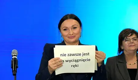 Monika Wielichowska na niebieskim tle trzymająca białą kartkę z czarnym napisem