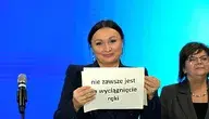 Monika Wielichowska na niebieskim tle trzymająca białą kartkę z czarnym napisem