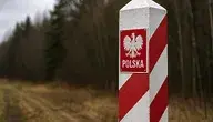 Słupek graniczny w barwach biało-czerwonych stojący na skraju lasu, symbolicznie oznaczający granicę Polski na tle rozmytego, leśnego krajobrazu.