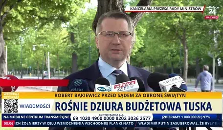 Mężczyzna, Przemysław Czarnek w garniturze udziela wypowiedzi mediom przed Kancelarią Prezesa Rady Ministrów w Warszawie. Na ekranie pasek informacyjny telewizji wPolsce24 dotyczący budżetu i polityki rządu Donalda Tuska