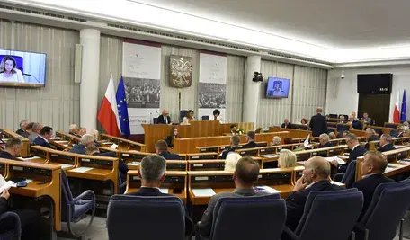 Wnętrze sali obrad Senatu Polski, widok na parlamentarzystów z flagą Polski i Unii Europejskiej, z udziałem osoby na ekranie wideo