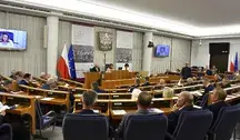 Wnętrze sali obrad Senatu Polski, widok na parlamentarzystów z flagą Polski i Unii Europejskiej, z udziałem osoby na ekranie wideo