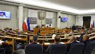 Wnętrze sali obrad Senatu Polski, widok na parlamentarzystów z flagą Polski i Unii Europejskiej, z udziałem osoby na ekranie wideo