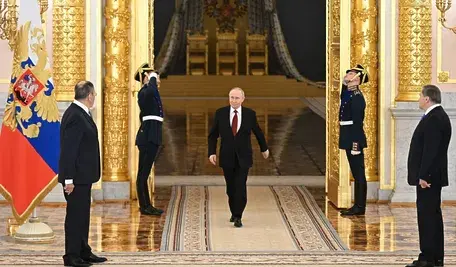 Władimir Putin przechodzi przez salę reprezentacyjną z dwoma wartownikami salutującymi, w tle złociste kolumny i rosyjska flaga państwowa