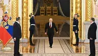 Władimir Putin przechodzi przez salę reprezentacyjną z dwoma wartownikami salutującymi, w tle złociste kolumny i rosyjska flaga państwowa