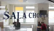 Widok przez szybę z napisem ‘SALA CHORYCH’ do sali szpitalnej, w której personel medyczny opiekuje się pacjentami w łóżkach