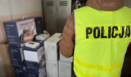 „Policjant w odblaskowej kamizelce z napisem POLICJA stoi obok stosu zapakowanych urządzeń RTV i AGD w kartonach, zabezpieczonych podczas akcji przeciwko grupie oszustów