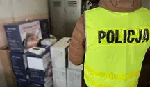 „Policjant w odblaskowej kamizelce z napisem POLICJA stoi obok stosu zapakowanych urządzeń RTV i AGD w kartonach, zabezpieczonych podczas akcji przeciwko grupie oszustów