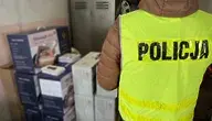 „Policjant w odblaskowej kamizelce z napisem POLICJA stoi obok stosu zapakowanych urządzeń RTV i AGD w kartonach, zabezpieczonych podczas akcji przeciwko grupie oszustów