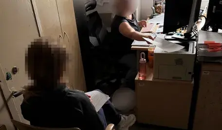 Zdjęcie przedstawia biuro, w którym przetwarzane są dokumenty. Dwie osoby siedzą przy biurku z komputerem i drukarką, jedna przekazuje dokumenty drugiej. Idealne do artykułów o śledztwach, zatrzymaniach, handlu zabytkami, sprawach kryminalnych w Polsce