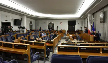 Sala Senatu RP z widokiem na miejsca senatorów, trybuny i sprzęt medialny; w tle flagi Polski i Unii Europejskiej oraz godło państwowe