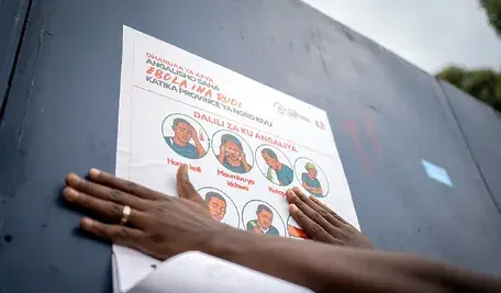 Edukacyjny plakat anty-Ebola w Demokratycznej Republice Konga, pokazujący objawy choroby i wskazówki ochronne; zdjęcie przedstawia osobę przyklejającą plakat w Gomie, Północne Kivu.