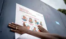 Edukacyjny plakat anty-Ebola w Demokratycznej Republice Konga, pokazujący objawy choroby i wskazówki ochronne; zdjęcie przedstawia osobę przyklejającą plakat w Gomie, Północne Kivu.
