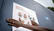Edukacyjny plakat anty-Ebola w Demokratycznej Republice Konga, pokazujący objawy choroby i wskazówki ochronne; zdjęcie przedstawia osobę przyklejającą plakat w Gomie, Północne Kivu.