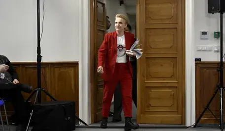 Joanna Scheuring-Wielgus wchodzi do sali konferencyjnej w czerwonym garniturze, trzymając dokumenty. Zdjęcie ilustruje jej udział w debacie publicznej w kontekście krytyki decyzji prezydenta Karola Nawrockiego o odebraniu Orderu Orła Białego Wołodymyrowi Zełenskiemu